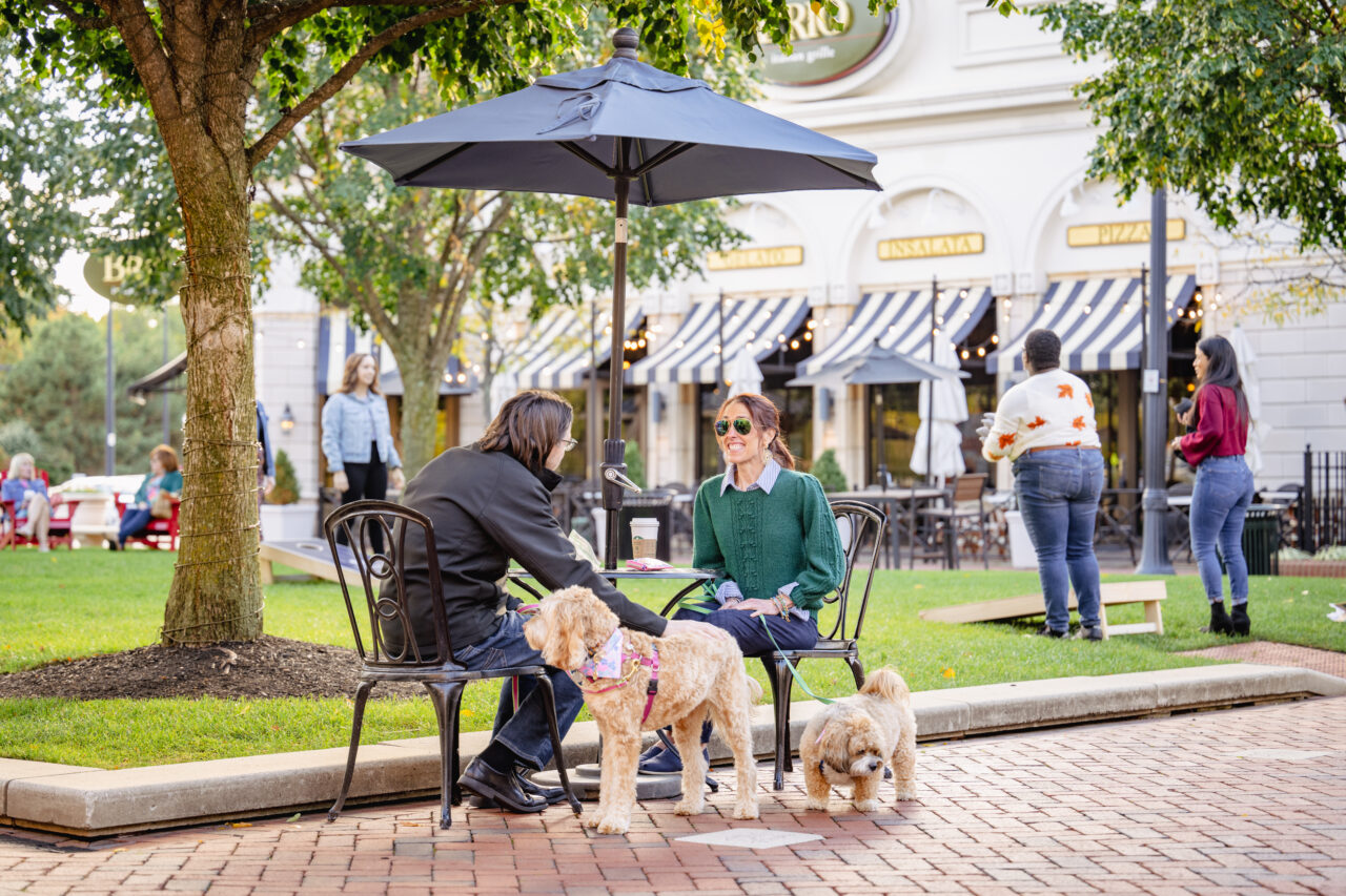 Two individuals sitting at a bistro table with their dogs outdoors at Legacy Village